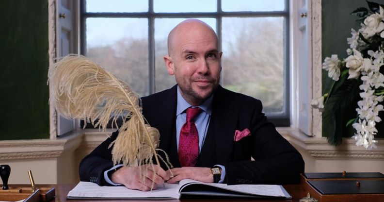 Tom Allen sat at a wooden desk, a window behind him, signing wedding documents with a very large quill.