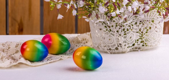 three rainbow easter eggs on a white table beside a white knitted pot with purple flowers