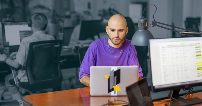 This is an image of a man working at a laptop computer. He is bald and is wearing a purple t shirt. He is in full colour and the rest of the image is in black and white.