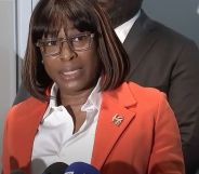 Philadelphia LGBTQ+ official Celena Morrison wears a white shirt and orange-red jacket as she speaks into microphones during a press conference