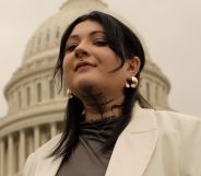 Non-binary activist Leah Juliett looks down at the camera while wearing a dark shirt and white blazer while standing in front of a US historic building