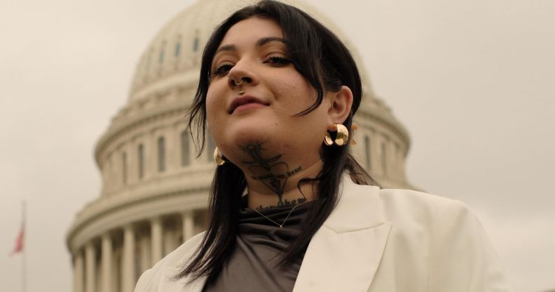 Non-binary activist Leah Juliett looks down at the camera while wearing a dark shirt and white blazer while standing in front of a US historic building