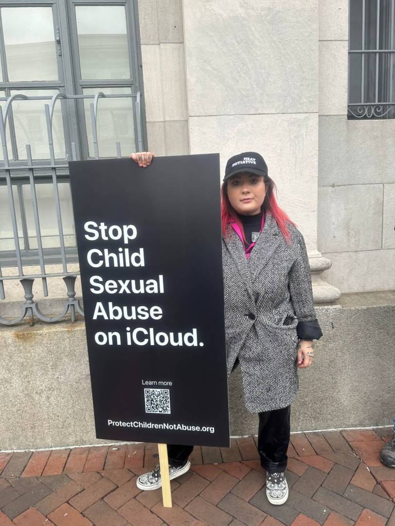Non-binary activist Leah Juliett holds a sign reading 'Stop child sexual abuse on iCloud' while wearing a grey coat and standing outside