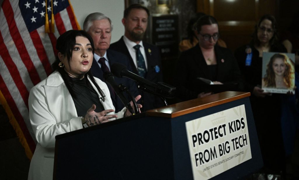Non-binary activist Leah Juliett wears a dark shirt and white jacket as they stand at a podium with a banner reading 'protect kids from big tech' during a press conference