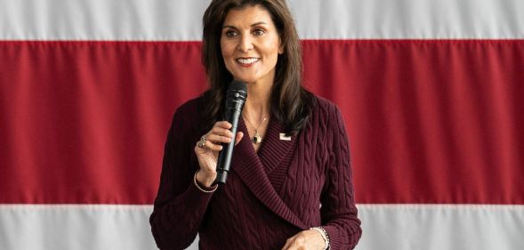 Republican politician Nikki Haley, who was running in the 2024 presidential election, speaks into a microphone while standing in front of the red and white stripes of the USA flag