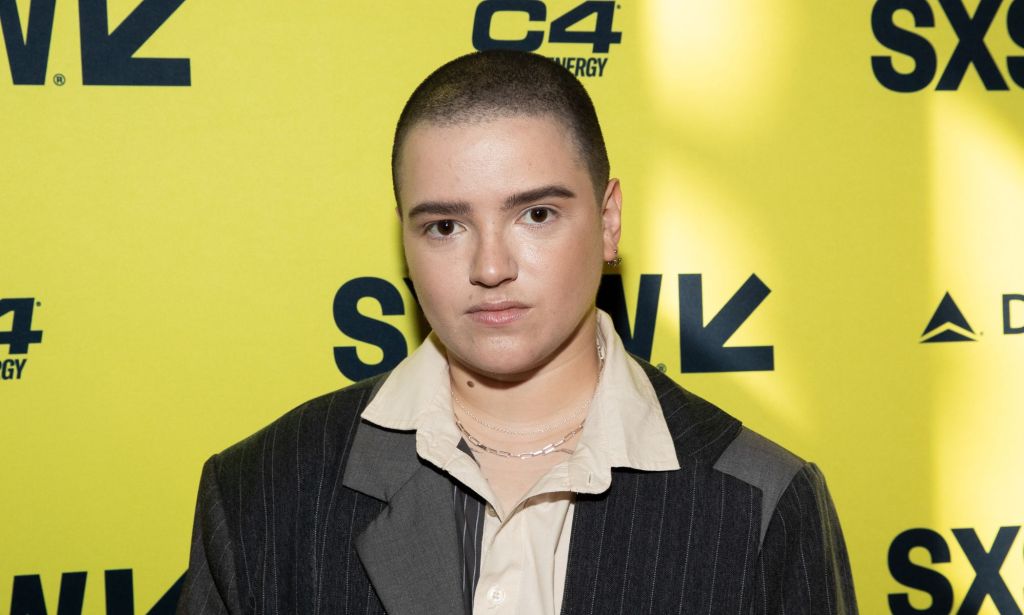 Star Trek: Discovery actor Blu del Barrio wears a grey and black blazer over a cream shirt while stood against a black and yellow background.