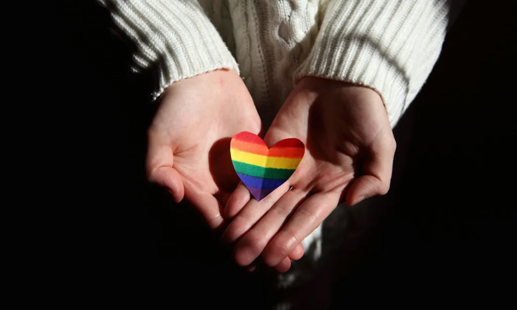 A person holds a rainbow striped heart in their hands towards the viewer