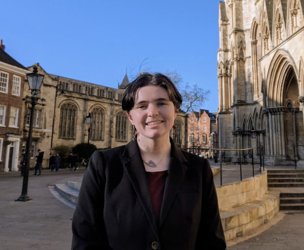 This is an image of a non-binary person standing outside in the courtyard, there is a cathedral style building in the background. They are wearing dark clothing and are smiling.