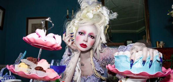 Bambie Thug in white face makeup and a towering blonde wig resting their hand on their chin.