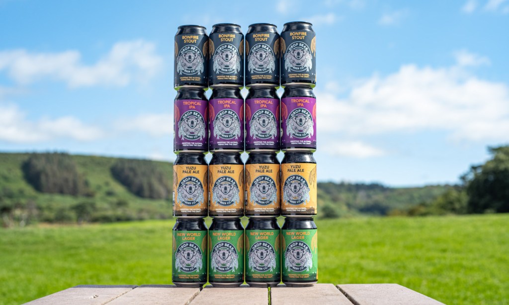 This is a product image of Drop Bear Beer Company. There are 4 columns of colourful cans on a table outside on a sunny day