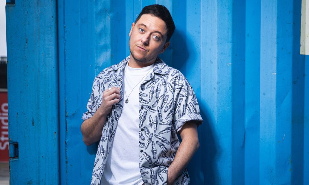 Emmerdale star and out trans actor Ash Palmisciano wears a white shirt and blue and grey patterned button up over it as he stares towards the camera