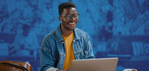 This is an image of a Black man smiling while looking down at his computer. He is wearing a denim shirt with a yellow tshirt underneath.