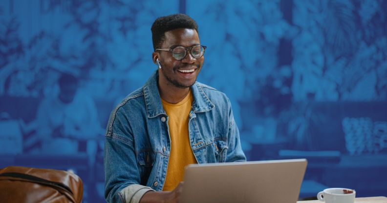 This is an image of a Black man smiling while looking down at his computer. He is wearing a denim shirt with a yellow tshirt underneath.