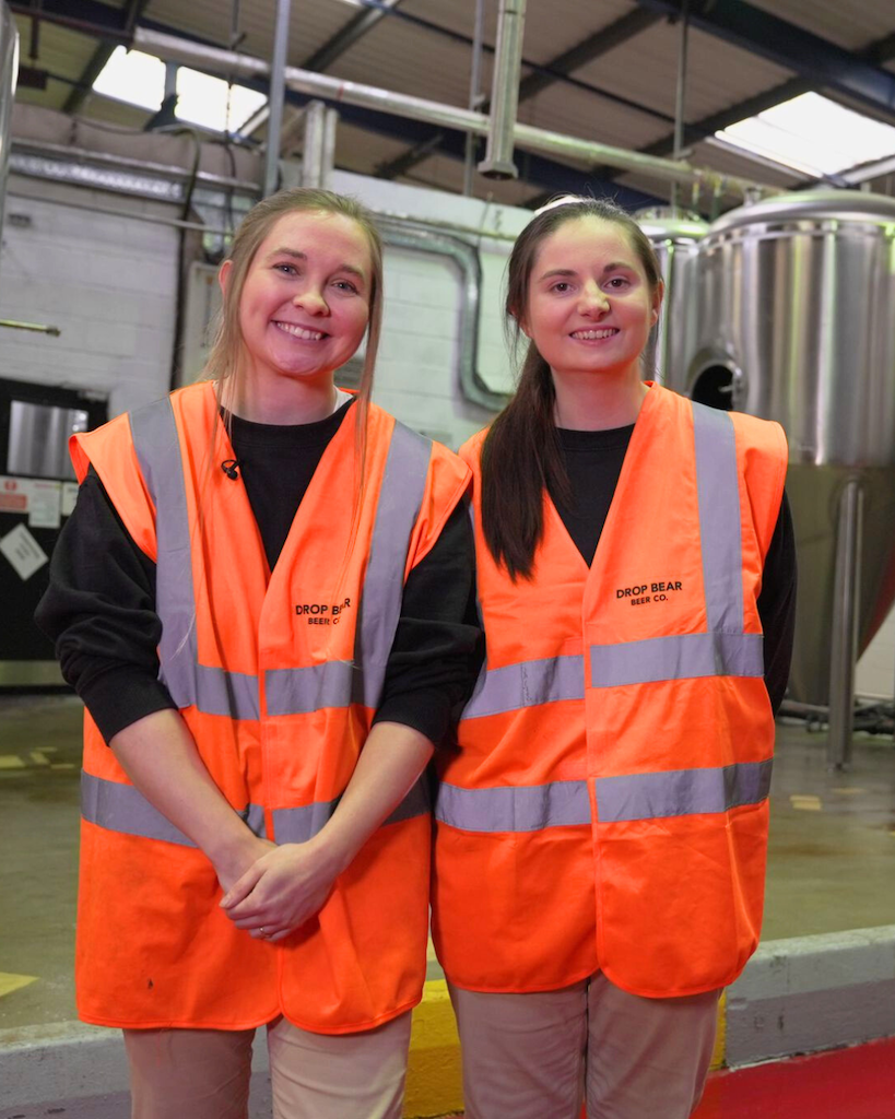 This is an image of two women standing in a brewery. they are both wearing black shirts with high visibility vests.