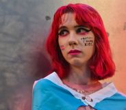 A person with red hair standing infront of a metal wall with paint that says "protect trans kids" on their cheek.