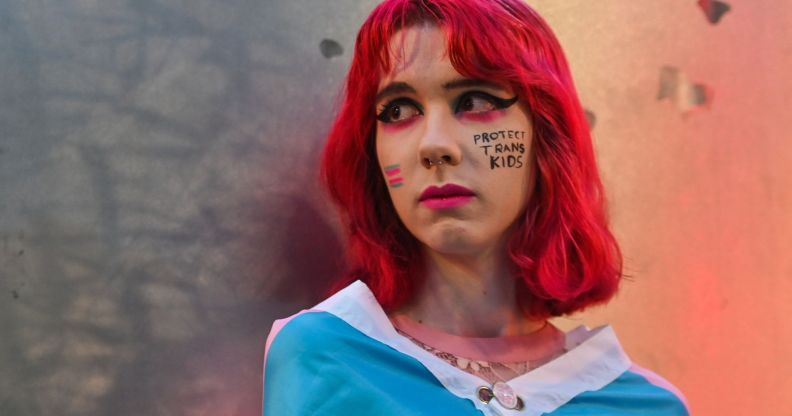 A person with red hair standing infront of a metal wall with paint that says "protect trans kids" on their cheek.