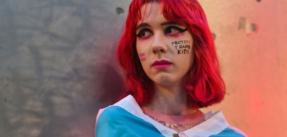 A person with red hair standing infront of a metal wall with paint that says "protect trans kids" on their cheek.