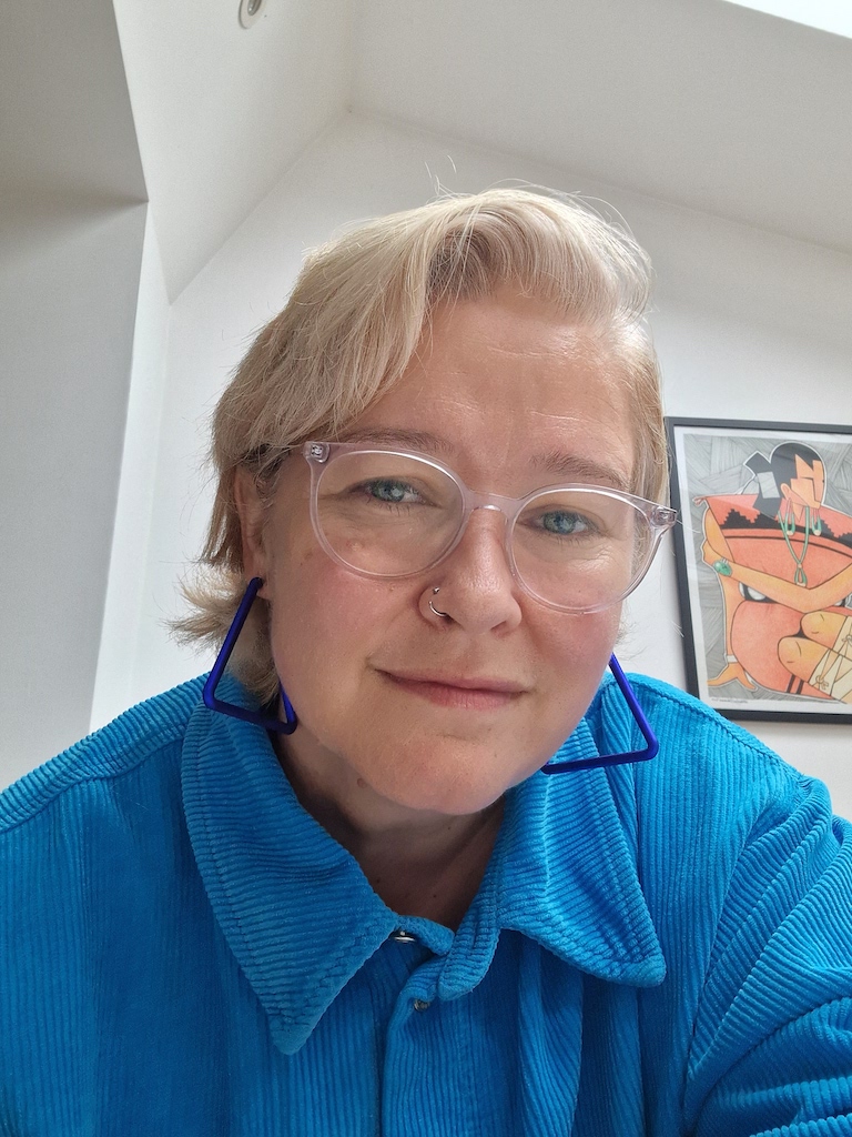 This is an image of a woman. She has shorter blonde hair and has clear framed glasses on. She is wearing a blue shirt with a high collar
