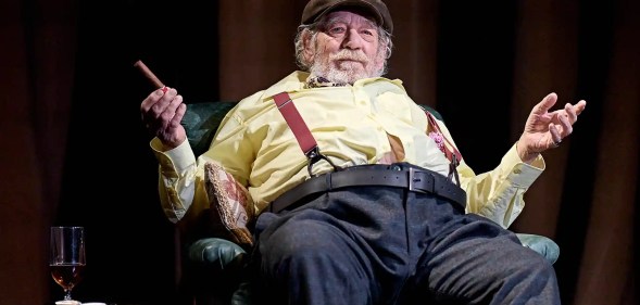 Ian McKellen in Player Kings, sitting on a chair, his chest and stomach fatter than usual, wearing a flat cap, yellow shirt and blue trousers, with red braces