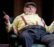 Ian McKellen in Player Kings, sitting on a chair, his chest and stomach fatter than usual, wearing a flat cap, yellow shirt and blue trousers, with red braces