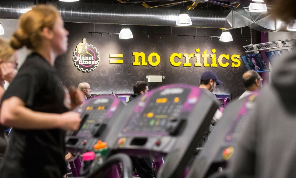 A person running on a treadmill in a Planet Fitness location. A mural on a wall says "no critics."