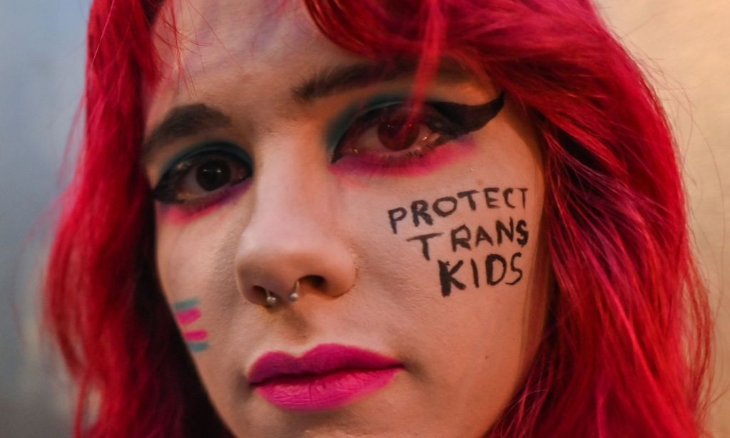 A close up of a trans activist with the words "protect trans kids" written on their face.