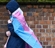 A hooded person walks past a brick wall with a trans flag around their back.