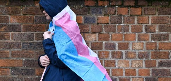 A hooded person walks past a brick wall with a trans flag around their back.