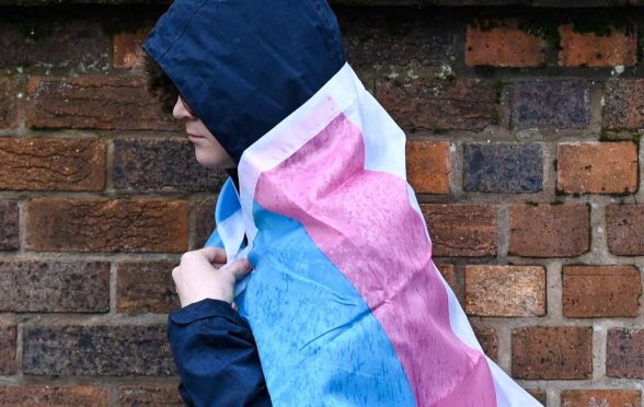 A hooded person wearing a trans flag for a cape in the rain.