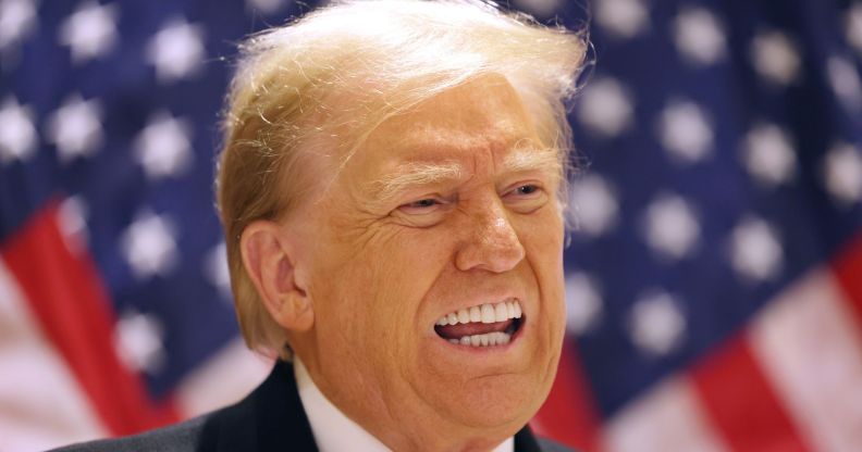 Former president Donald Trump wears a suit and tie as he speaks to people off camera and stands in front of red, white and blue US flags