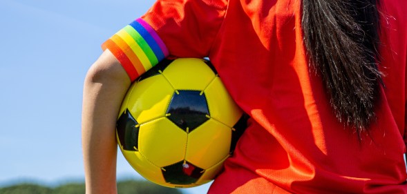 Individual in a orange sports top holding a football under their arm with a rainbow armband