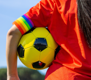 Individual in a orange sports top holding a football under their arm with a rainbow armband