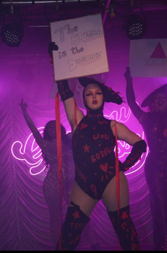 Kyle Cook, a drag queen and activist living with HIV, wears an HIV-inspired drag outfit while holding up a sign slamming stigma about the diagnosis 