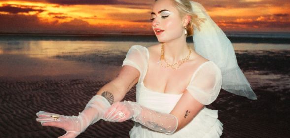 Låpsley stands on a beach during the sunset wearing a wedding dress and veil. She is holding out her gloved hand.