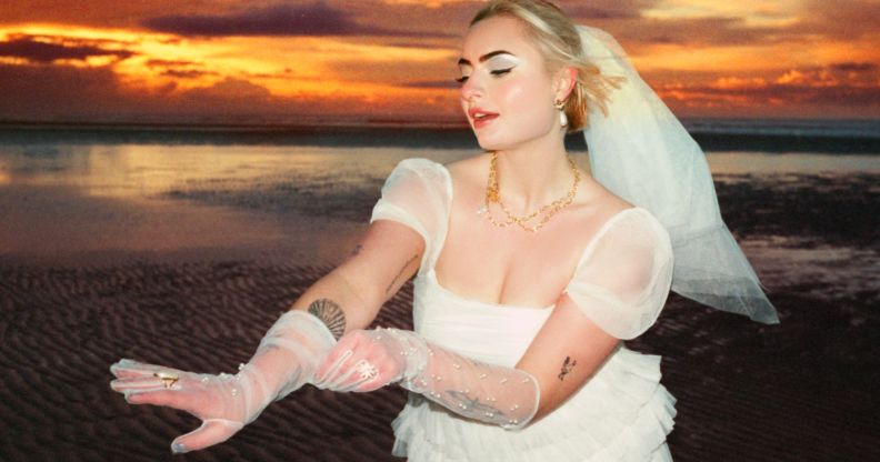 Låpsley stands on a beach during the sunset wearing a wedding dress and veil. She is holding out her gloved hand.