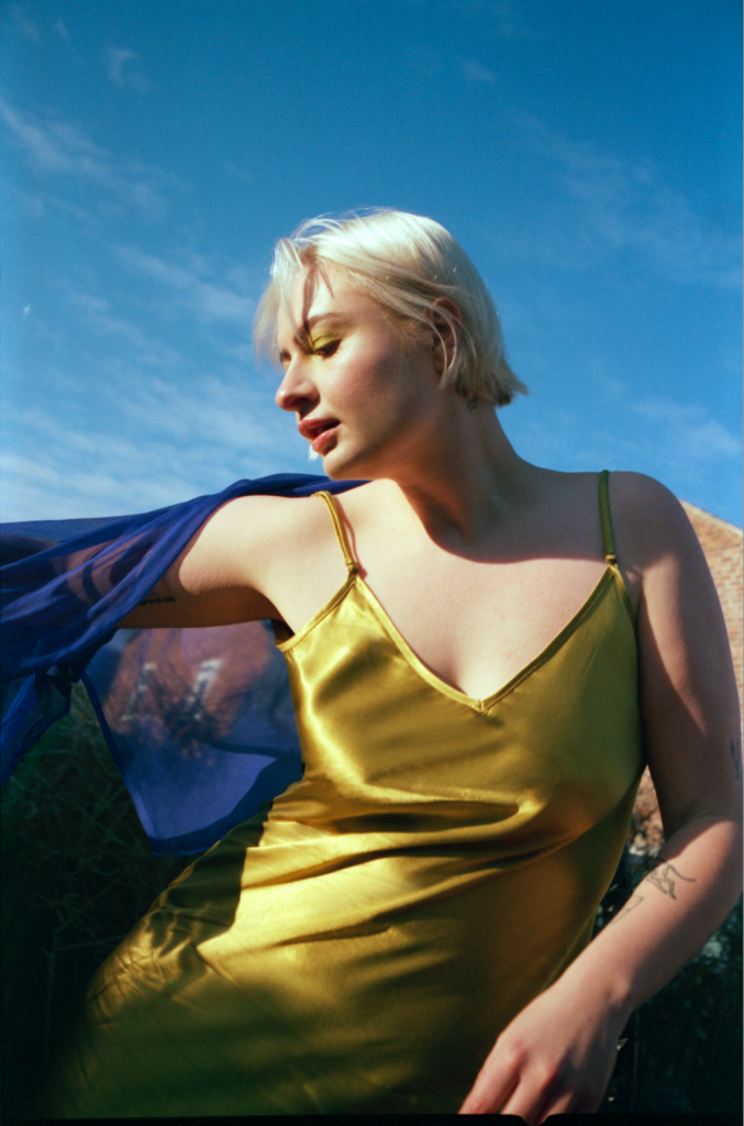 Låpsley in a yellow sleeveless dress. She is outside and the sky is blue behind her. She is looking away from the camera and down at the ground.