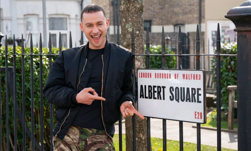 olly Alexander smiles and points at the EastEnders Albert Square sign.