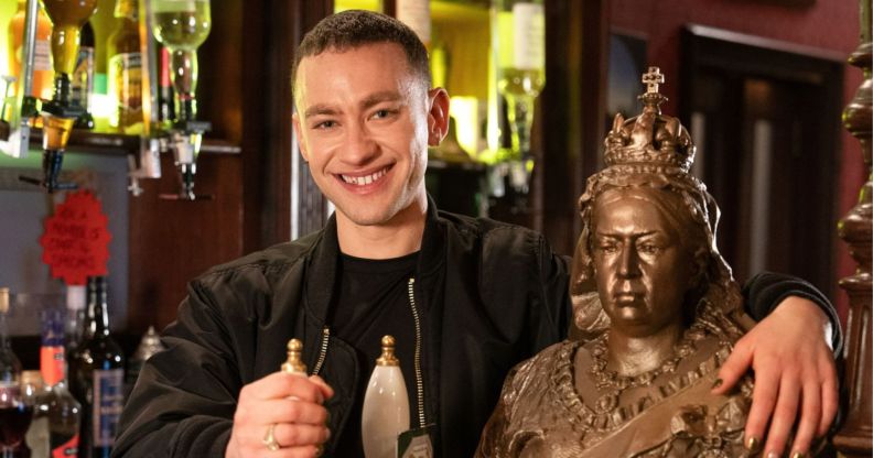 Olly Alexander smiles as he stands behind the bar in EastEnders pub The Queen Vic, pulling a pint.