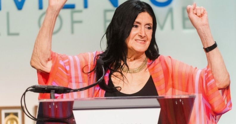 Trans academic and activist Sandy Stone wears a dark shirt and pink top as she holds up her hands while being inducted into the US National Women's Hall of Fame
