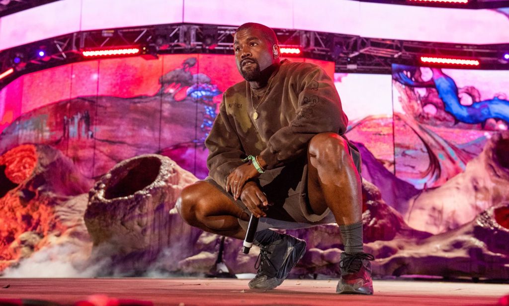 Rapper Ye, formerly known as Kanye West, wears a brown shirt and shorts as he crouches on stage during a performance