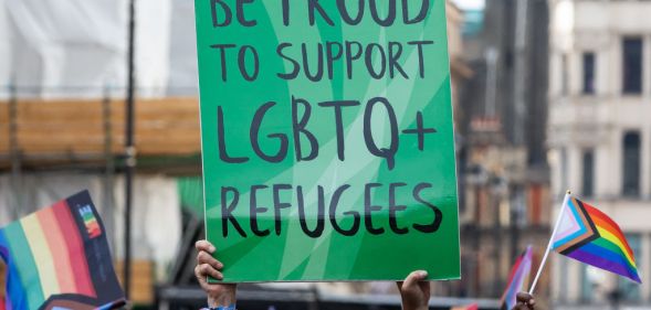 This is an image of a sign in black text on a green board. The text reads "Be Proud to support LGBTQ+ refugees". THere are various pride flags in hand