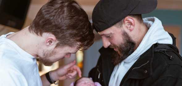Two caucasian men in their thirties are affectionally holding and looking at a newborn baby lovingly.