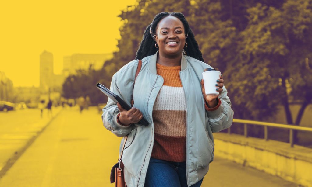This is an image of a Black woman walking to work. She has a coffee in one hand and holding a laptop computer in the other. 