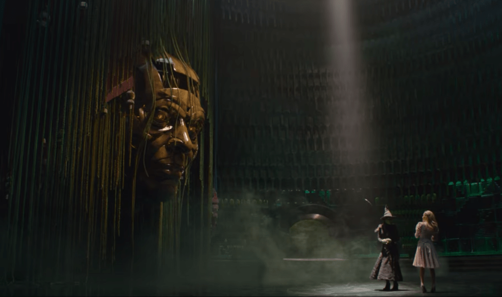 A huge puppet head appears from some curtains in Emerald City, dwarfing witches Elphaba and Gilnda who watch on