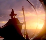 Screenshot from Wicked trailer showing Elphaba in her trademark pointed witch hat and cape and broomstick, about to leap from a broken window. The sun is setting behind her