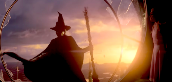 Screenshot from Wicked trailer showing Elphaba in her trademark pointed witch hat and cape and broomstick, about to leap from a broken window. The sun is setting behind her