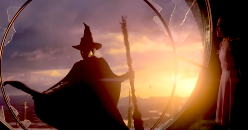 Screenshot from Wicked trailer showing Elphaba in her trademark pointed witch hat and cape and broomstick, about to leap from a broken window. The sun is setting behind her