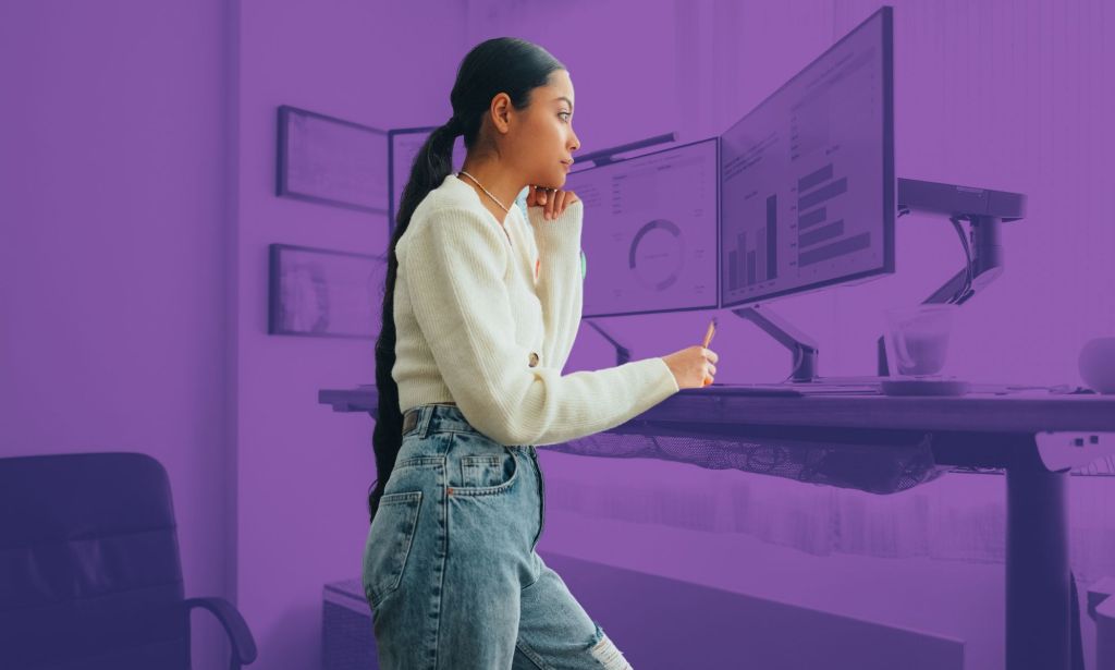 This is an image of a woman standing at a desk looking at computer monitors.