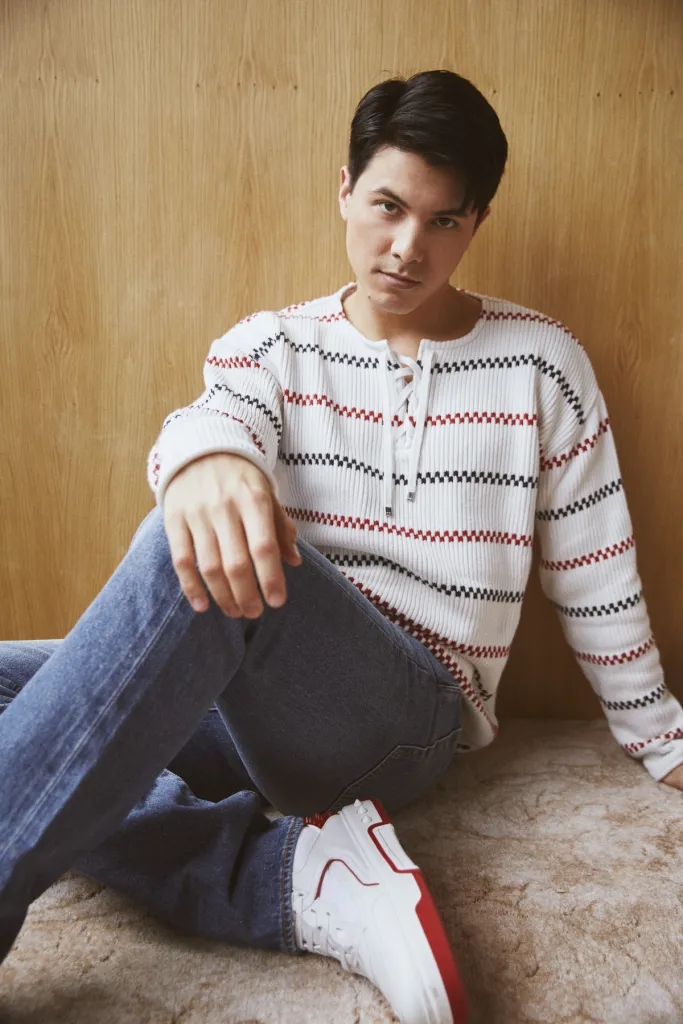 James Phoon poses while sitting against a brown wall and wearing blue jeans white and red trainers and a white striped jumper.