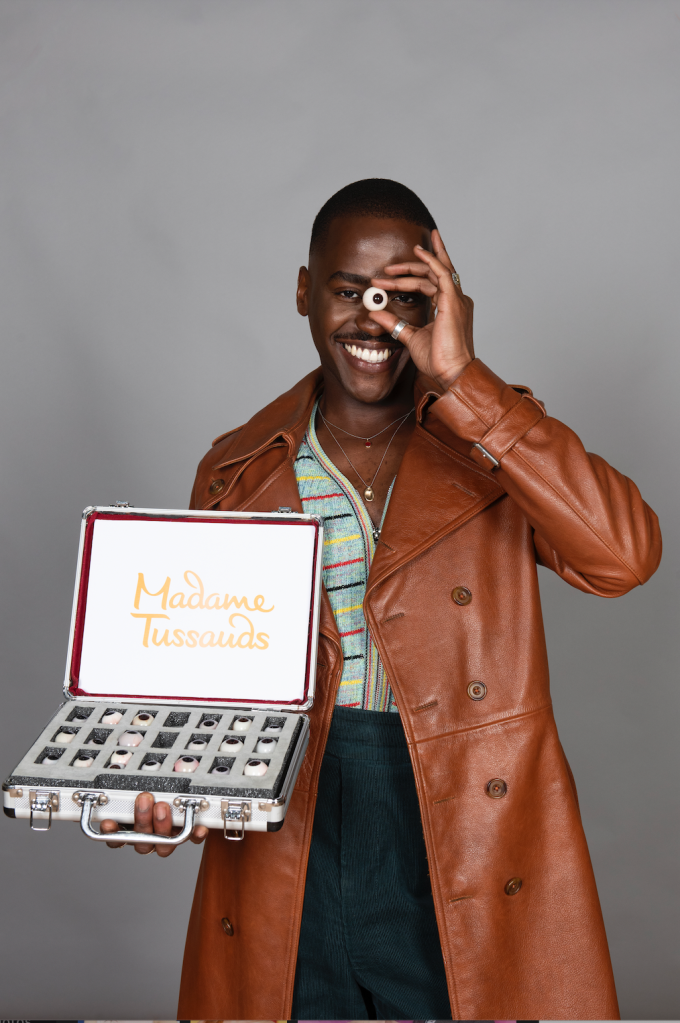 Doctor Who star Ncuti Gatwa in a brown coat and colourful cardigan holding a box of fake eyes and an eye up to his face.
